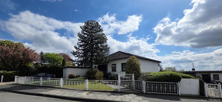 Ein einstöckiges Wohnhaus mit weißem Zaun und gepflegtem Garten unter einem weiten, wolkenverhangenen Himmel.